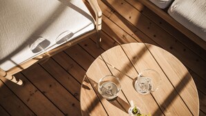 Ein runder Holztisch mit zwei Gläsern und einer Flasche, umgeben von Holzdielen und einem Stuhl mit weißem Sitzkissen.