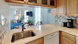 A kitchen with wooden cabinets, a stainless steel sink, and a dishwasher. There is a dining area with a blue sofa and a television in the background. - Dolphin's Den | Ocean Views + Community Pools (North Topsail Beach)