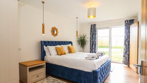 A bedroom with a large bed, a bedside table, a hanging light fixture, and a view of the outdoors through a sliding door. - Polly's Annexe (Ipswich)