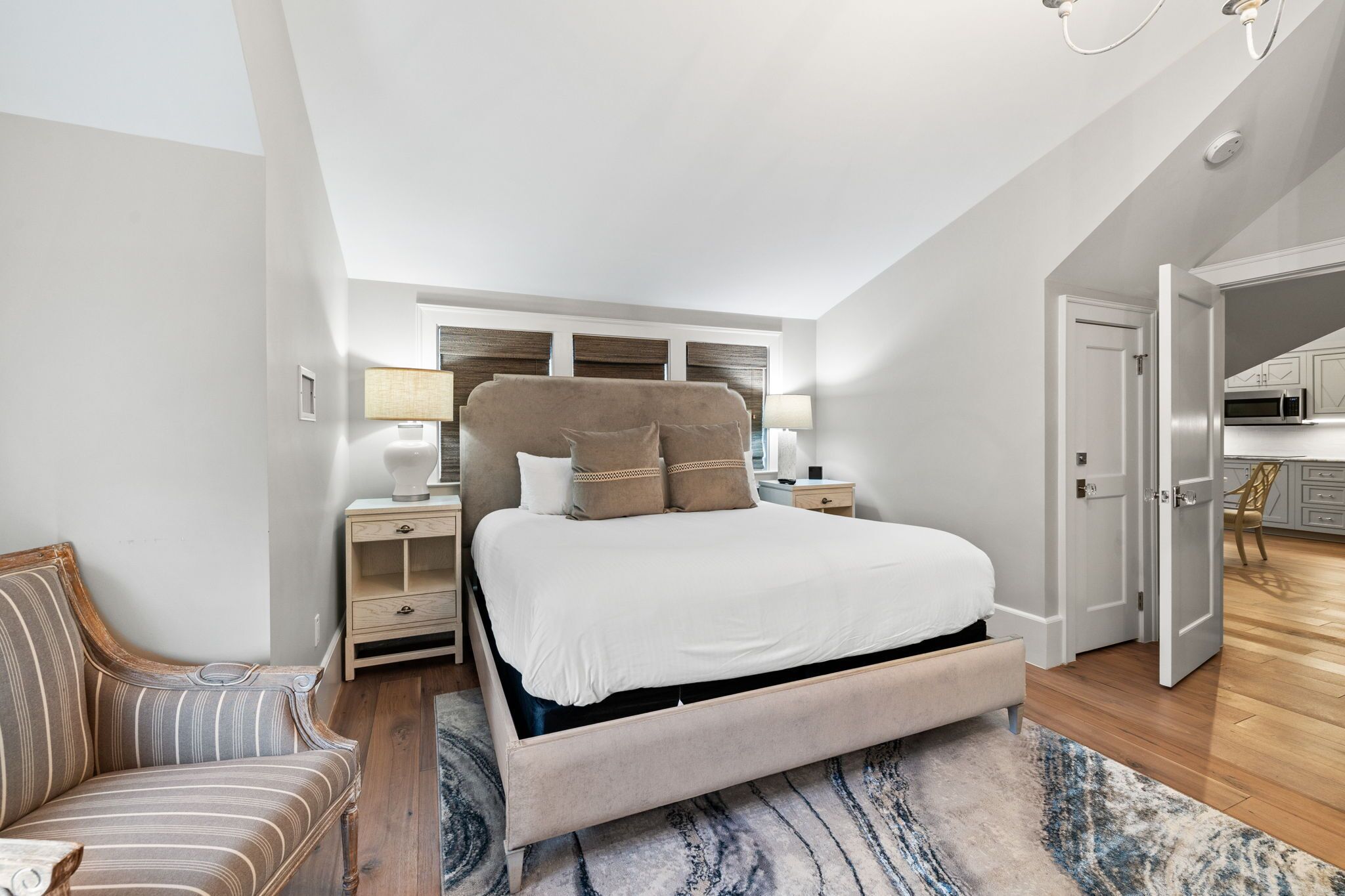 A bedroom with a large bed, a nightstand, a lamp, a chair, and a view of the kitchen.