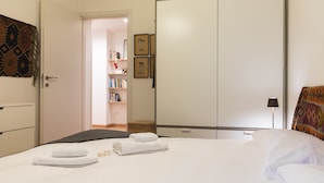 A neatly made bed with white linens and two folded towels, a bookshelf with books and framed pictures, and a door leading to another room. - notaMi - Dimora Certosa (Milan)