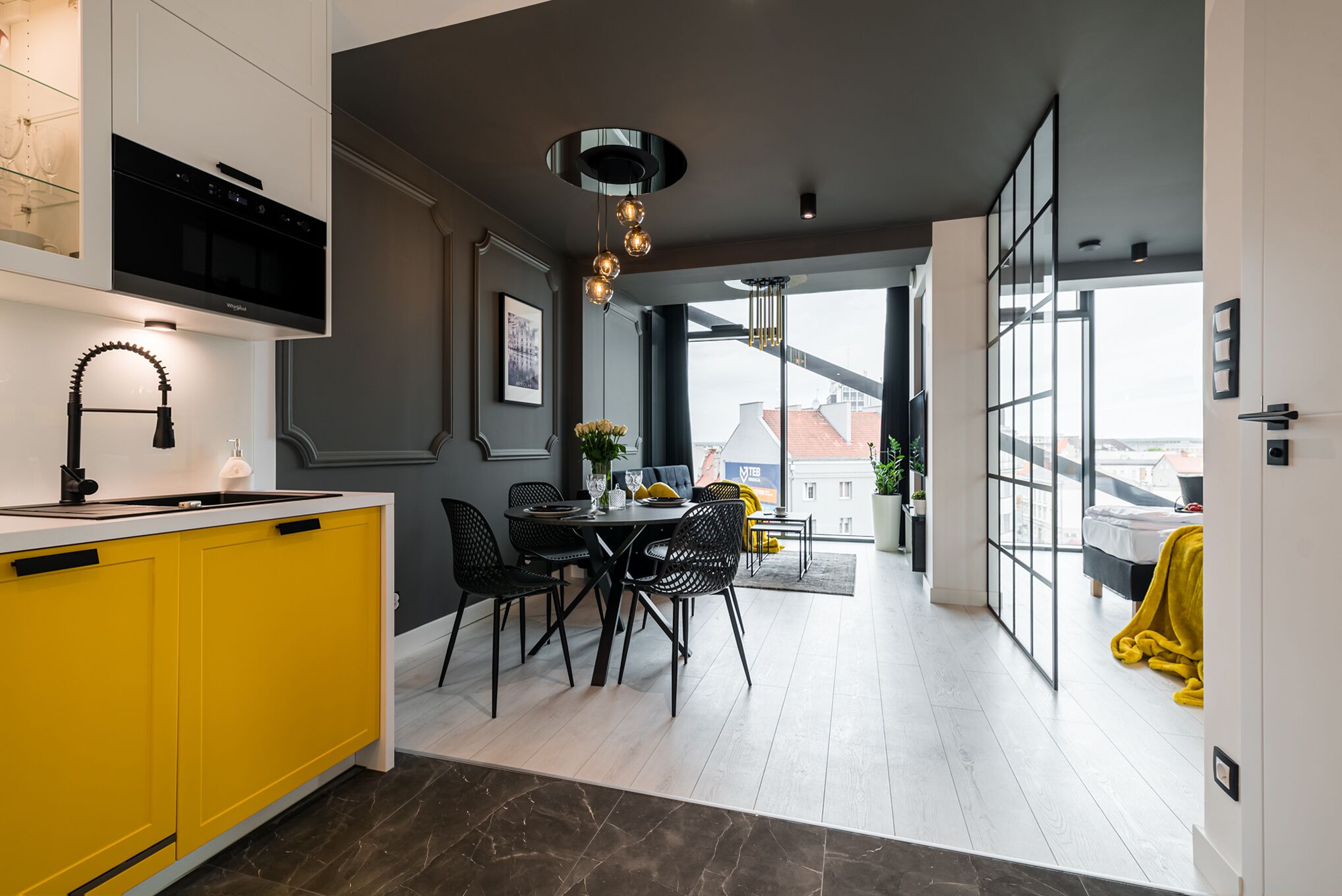 Een moderne keuken met gele kastjes, een zwarte afzuigkap, en een eethoek met een ronde tafel en zwarte stoelen.