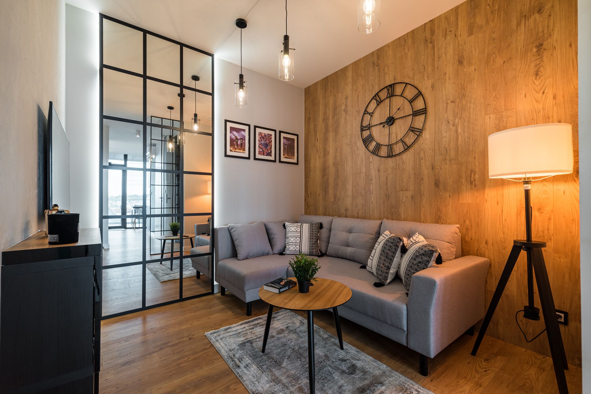 A modern living room with a grey sofa, a round wooden coffee table, a large wall clock, and a floor lamp.
