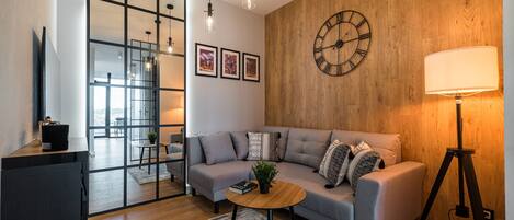 A modern living room with a grey sofa, a round wooden coffee table, a large wall clock, and a floor lamp.