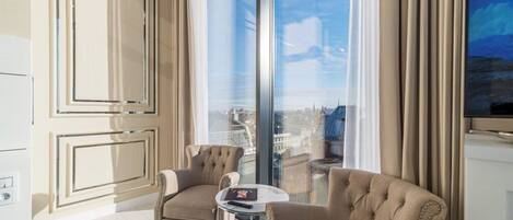 A modern hotel room with two plush armchairs, a small round table, and a large window offering a city view.