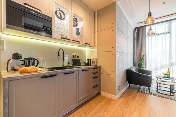 A modern kitchen with a built-in oven, a sink, and a countertop with a loaf of bread. There is a black chair and a small round table with a plant.