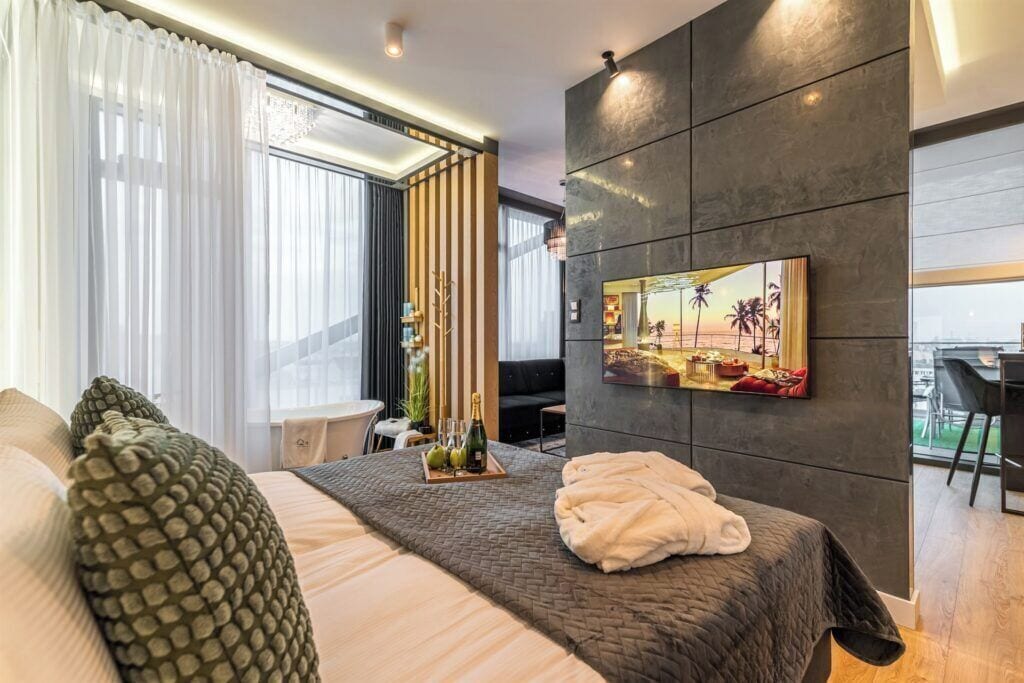 Camera d'albergo moderna con un letto grande, una TV a schermo piatto montata a parete, un'area pranzo con tavolo e sedie e vista sulla città attraverso ampie finestre.