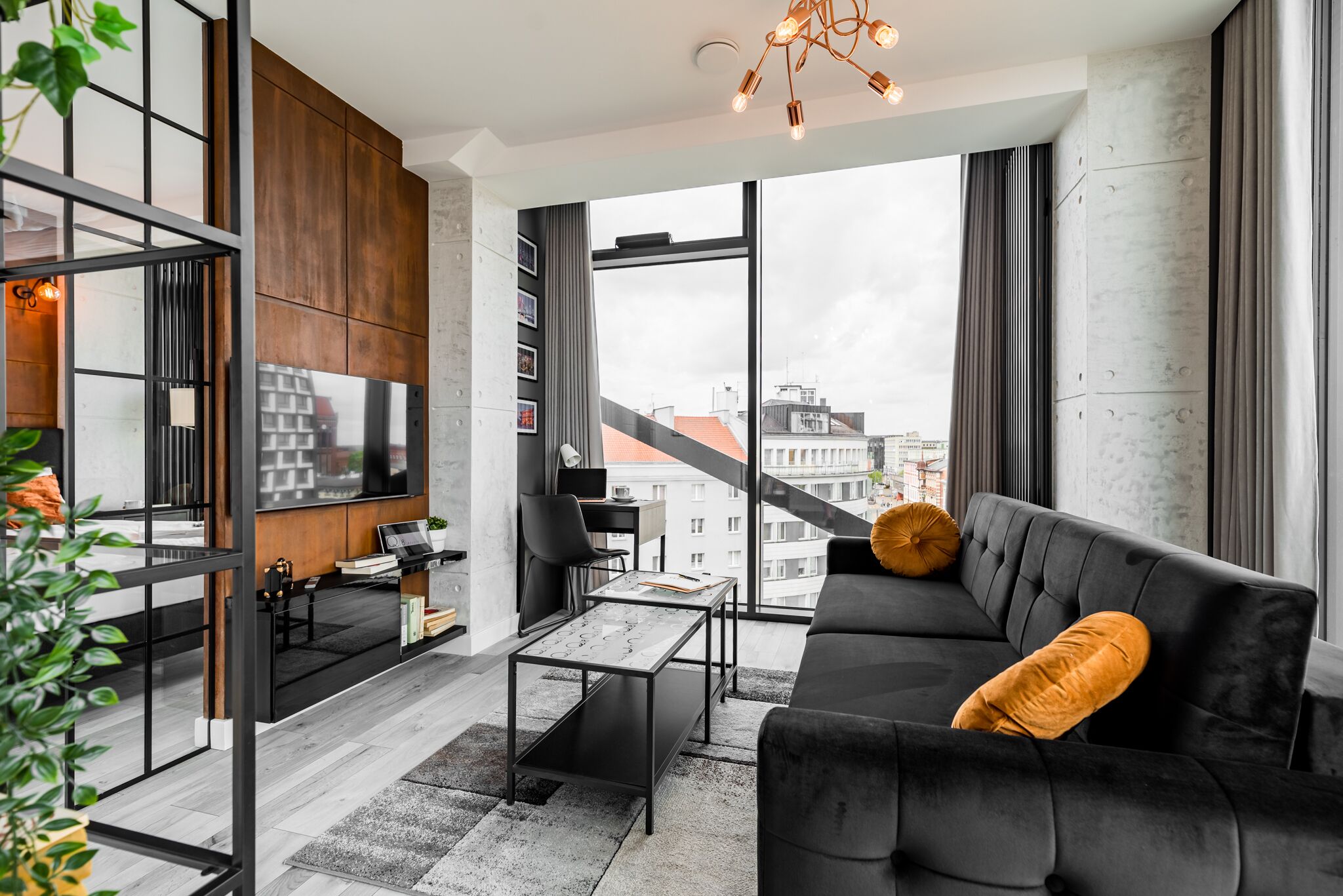 A modern living room with a black sofa, a coffee table, and a TV. There is a large window with a view of buildings outside.
