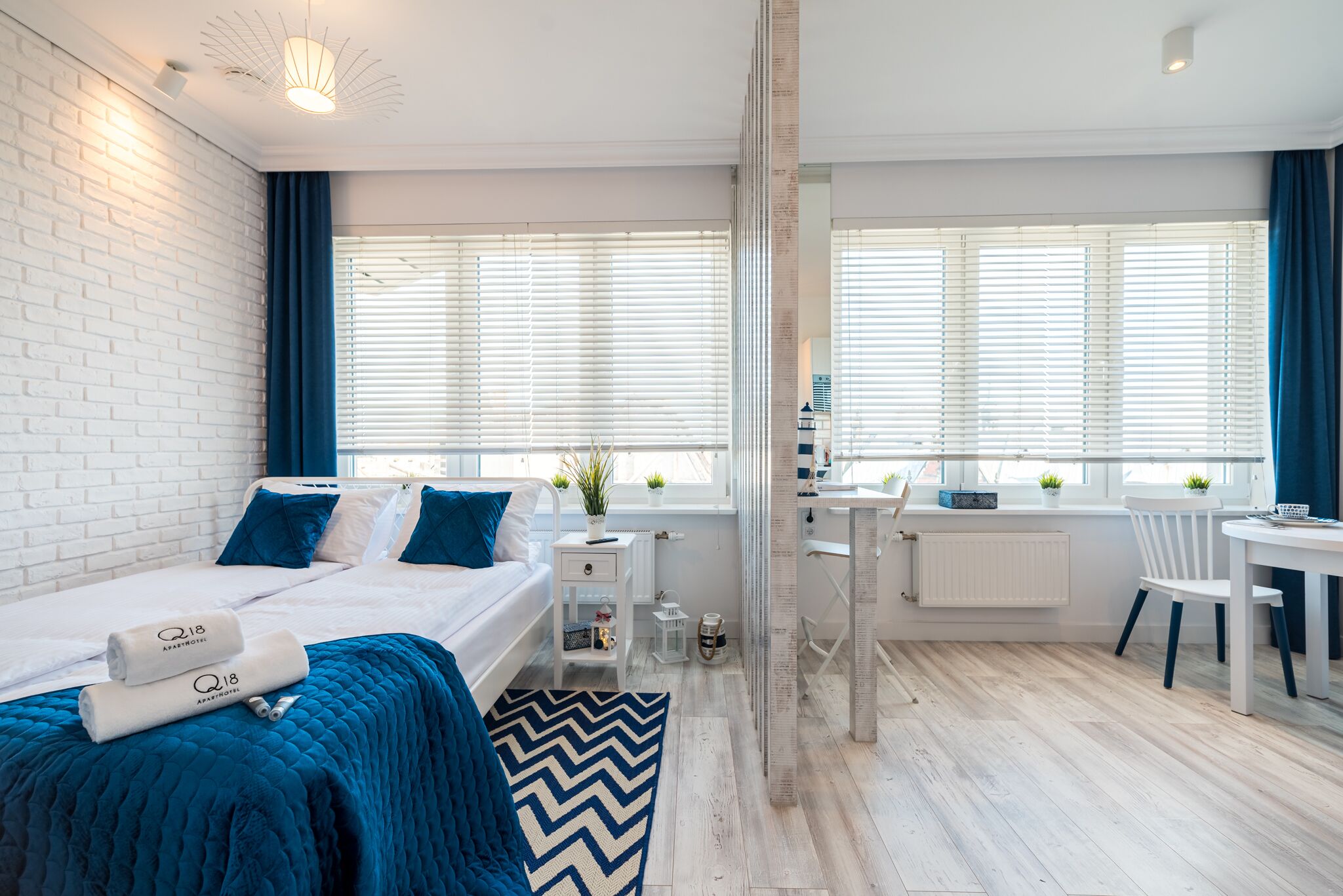 A modern hotel room with a white brick wall, a bed with blue bedding, a desk with a chair, and a view through a glass door.
