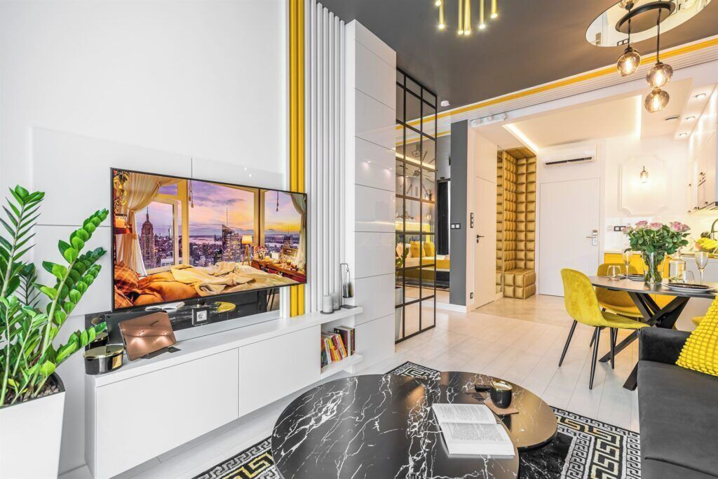 Un salon moderne doté d’un grand téléviseur à écran plat, d’un coin repas avec des chaises jaunes et d’une cuisine visible à travers une porte ouverte.