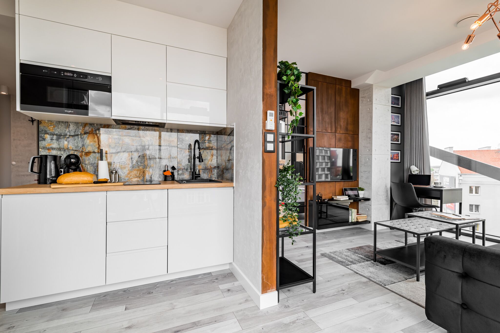 A modern kitchen with white cabinets, a built-in oven, and a marble backsplash. There is a wooden column with a plant, and a dining area with a black sofa and a table.