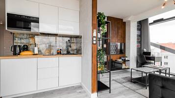 A modern kitchen with white cabinets, a built-in oven, and a marble backsplash. There is a wooden column with a plant, and a dining area with a black sofa and a table.