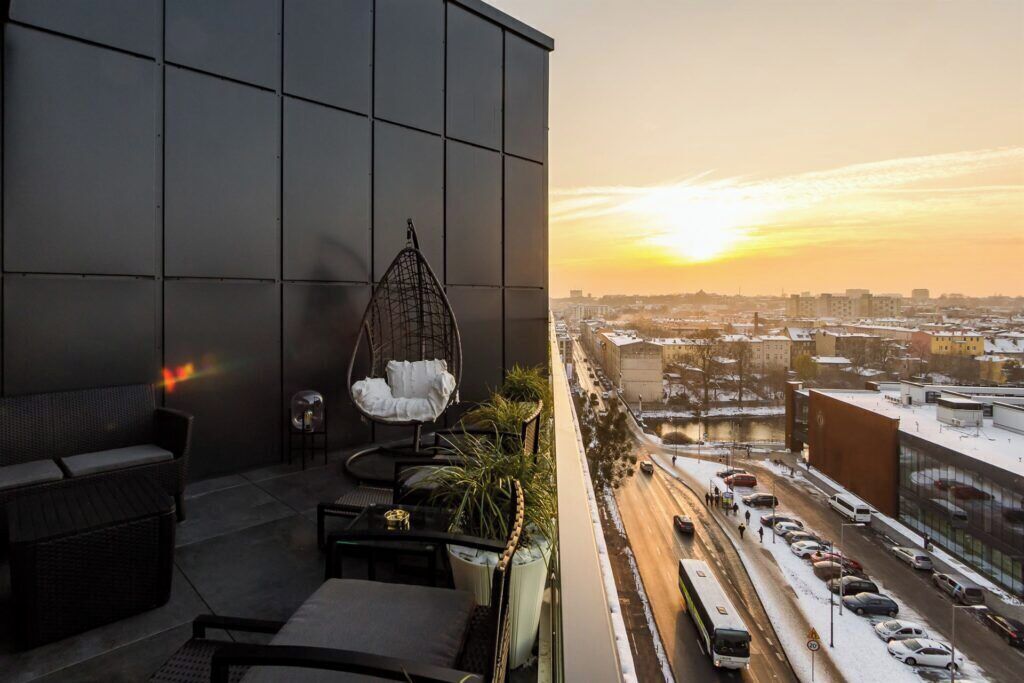 A balcony with a hanging chair, a sofa, and a table, overlooking a cityscape with a sunset.