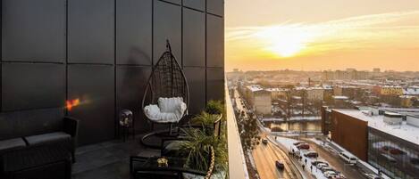 A balcony with a hanging chair, a sofa, and a table, overlooking a cityscape with a sunset.