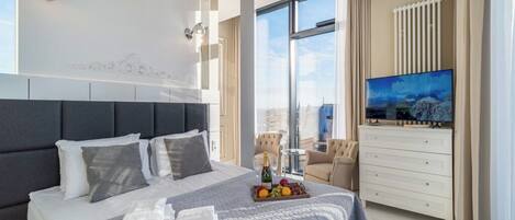 A modern hotel room with a large bed, a flat-screen TV, a dining area with a table and chairs, and a view of the city through large windows.