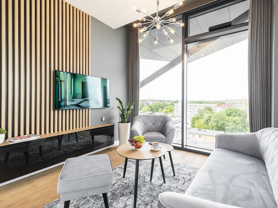 Un salon moderne avec une grande télévision, un canapé gris, une petite table sur laquelle se trouve un saladier rempli de fruits, et une vue sur la ville grâce à de grandes fenêtres.