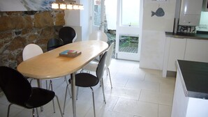A dining area with a wooden table and black chairs, a stone wall with a painting, a kitchen with white cabinets and a refrigerator, and a door leading to a balcony.