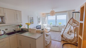A modern kitchen with white cabinets, a sink, and a dining area with a white chair and a hanging chair. A large window offers a view of the sea. - 13 Carrack Widden - amazing sea views with spa passes (St Ives)