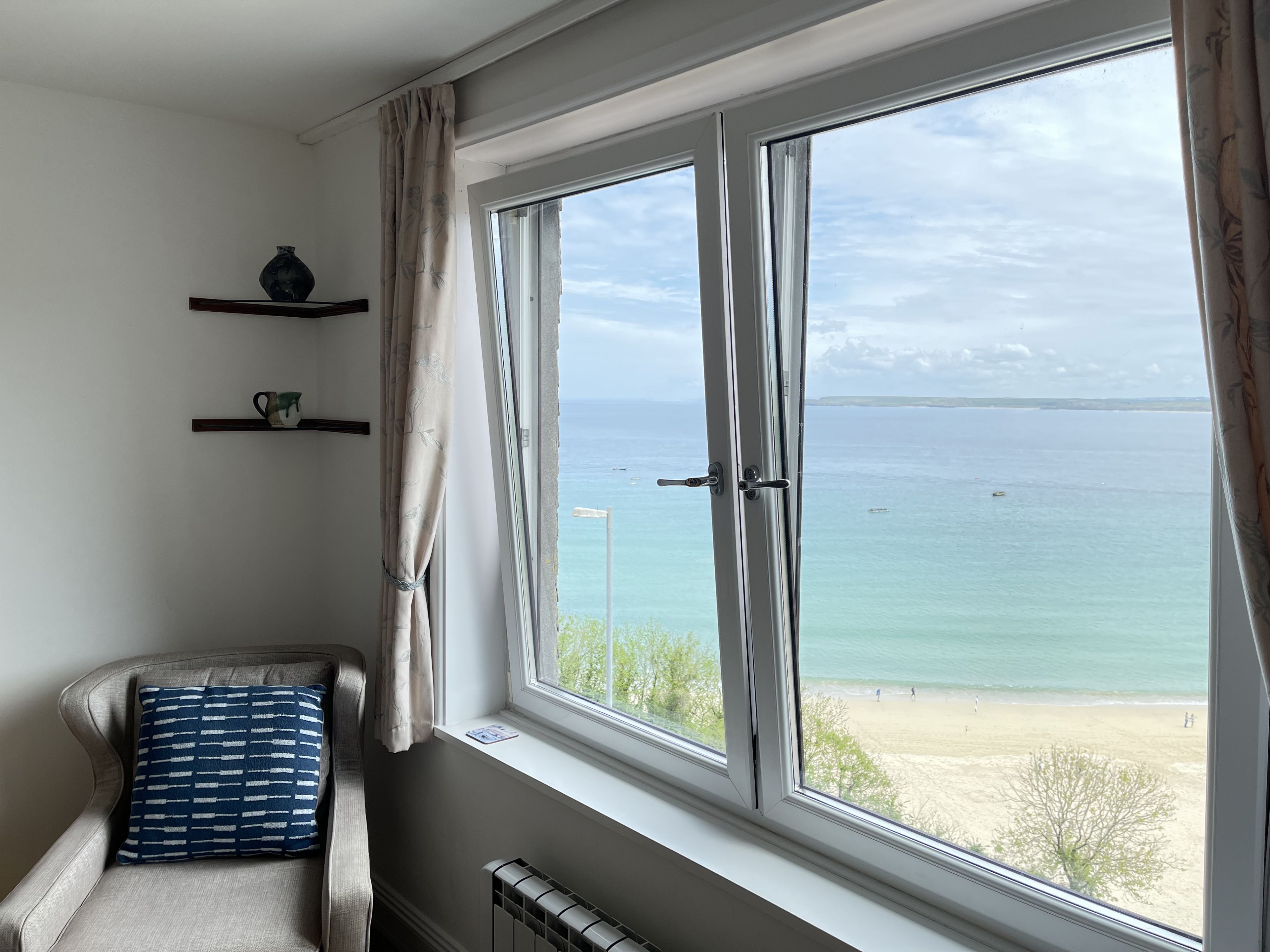A room with a large window overlooking a beach, a radiator, a chair with a patterned cushion, and two shelves with decorative items.