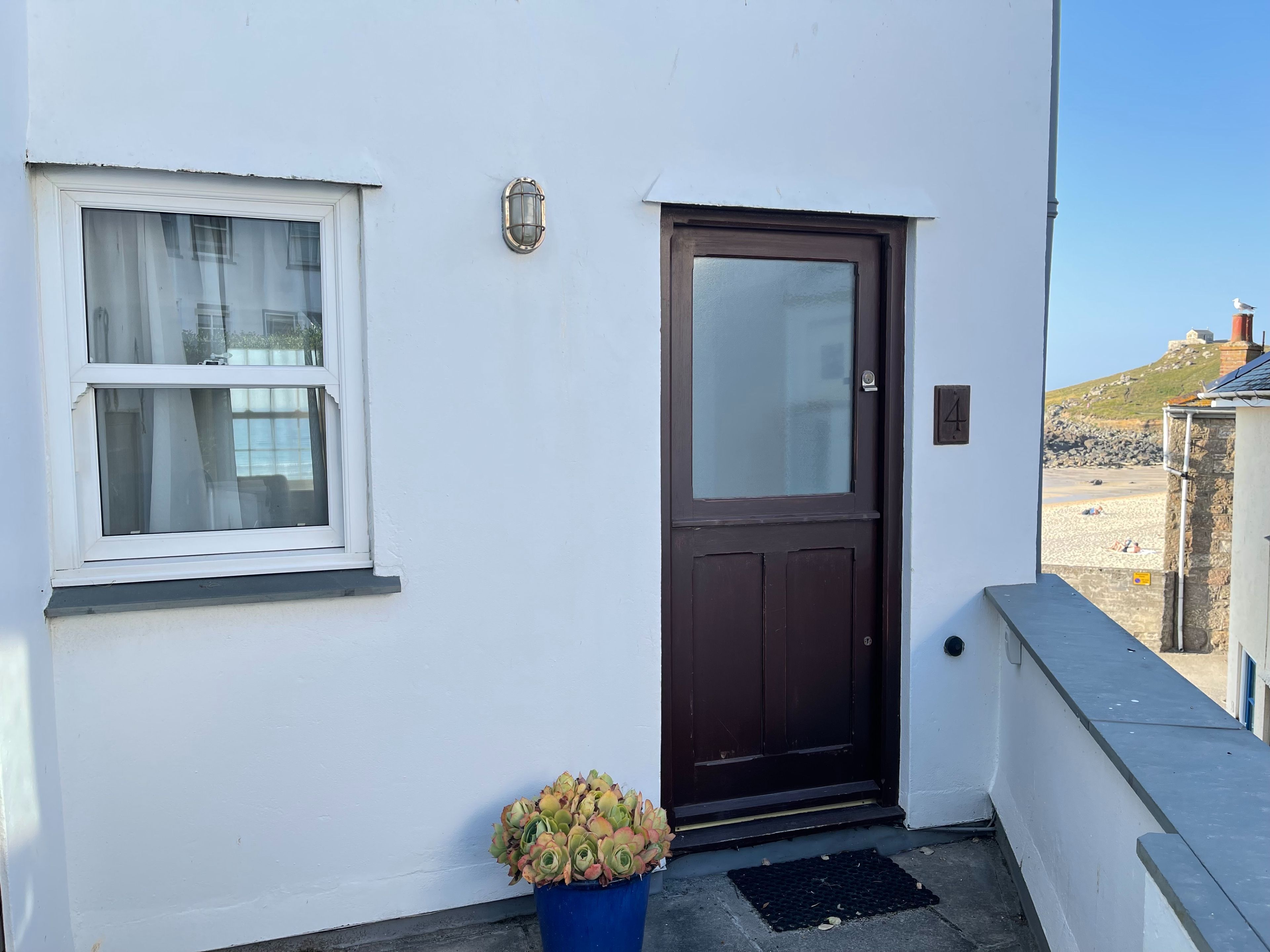 A white building with a brown door, a window with a white frame, and a blue pot with plants.