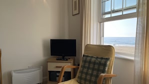 Et værelse med fjernsyn på et træskab, en stol med ternet pude, en radiator og et vindue med udsigt til stranden.