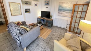 Una sala de estar con un sofá gris, un sillón blanco, una mesa de centro y un televisor sobre un mueble de madera. Hay cuadros enmarcados en las paredes, una ventana y una puerta con panel de vidrio.