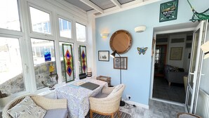Una habitación iluminada por el sol con muebles de mimbre, una mesa con mantel estampado y una pared con un cuadro enmarco y un espejo decorativo.