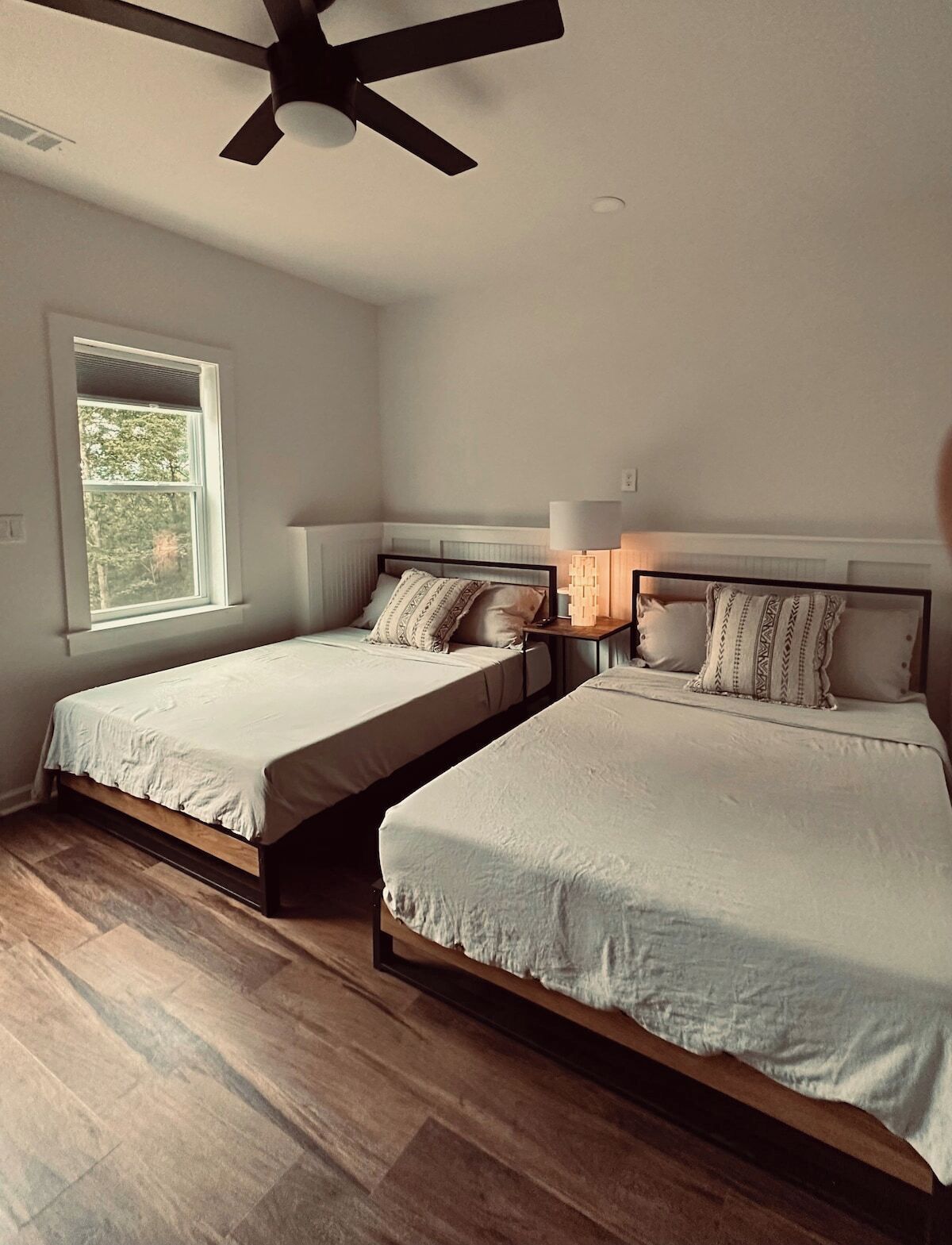 Une chambre avec deux lits, un ventilateur de plafond, une fenêtre et une table de chevet avec une lampe.
