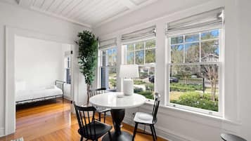 A room with a round dining table, two chairs, a potted plant, and a view of the outdoors through large windows.
