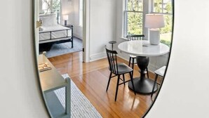 Een kamer met een ronde tafel en stoelen, een bed met een wit hoofdeinde, en een raam met uitzicht op groen.
