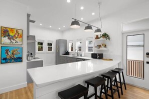A modern kitchen with white cabinetry, a large island with bar stools, pendant lighting, and artwork on the walls. - Otis Blue at Angels Quarter | Home Steps to 7th! (Tampa)