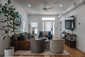 A spacious living room with a ceiling fan, a large potted plant, a sofa set, a coffee table, and a TV on a wooden wall. - Otis Blue at Angels Quarter | Home Steps to 7th! (Tampa)