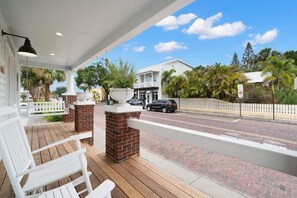 Een overdekte veranda met witte stoelen, een bakstenen pilaar en uitzicht op een straat met auto's en een witte hek.