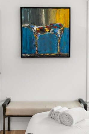 Una cama con sábanas blancas y una toalla blanca enrollada, un banco y un cuadro abstracto colorido en la pared.