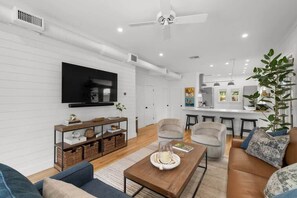 Uma sala de estar moderna com televisão, sofás, uma mesa de centro e uma cozinha ao fundo.
