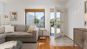 A modern living room with a grey sofa, a round coffee table, and a small side table. A large window with a view of buildings and a door leading outside are visible.
