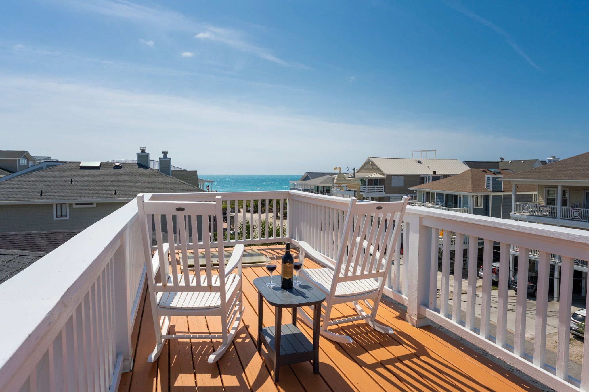 Una terraza de madera con barandillas blancas, dos sillas mecedoras blancas y una mesita con una botella y dos copas de vino. La terraza da a una playa con casas al fondo.