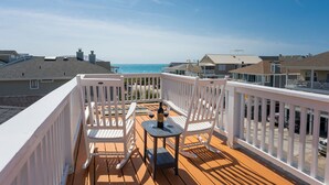 A wooden deck with white railings, two white rocking chairs, and a small table with a bottle and two wine glasses. The deck overlooks a beach with houses in the background.
