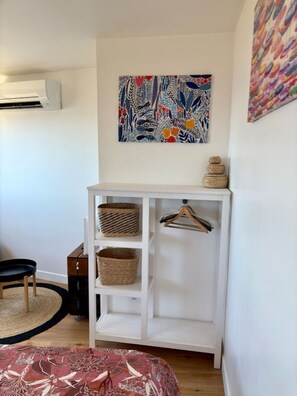 A bedroom with a white wardrobe, a hanging basket, a chair, and a colorful abstract painting on the wall.