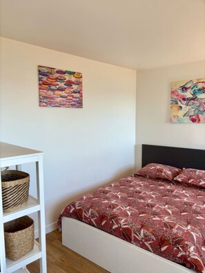 Un dormitorio con una cama, una estantería blanca con cestas y dos cuadros en la pared.