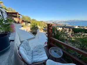 A balcony with a wicker chair and a small table, overlooking a coastal landscape with buildings and a clear sky.