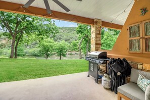 Een overdekt terras met een barbecue, een bank en uitzicht op een rivier en bomen.