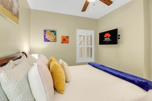 Een slaapkamer met een bed, kussens, een televisie, een plafondventilator en een raam met zonwering.