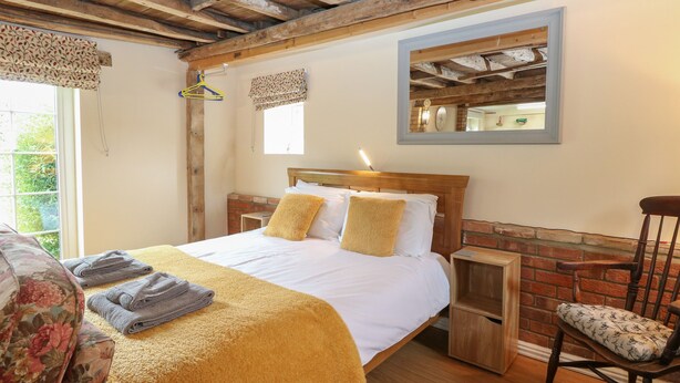 Ein Schlafzimmer mit Holzboden, einer Ziegelwand, einem Bett mit weißer Bettwäsche und zwei Kissen, einem Nachttisch aus Holz, einem Stuhl mit gemustertem Sitzkissen und einem Fenster mit Vorhang.