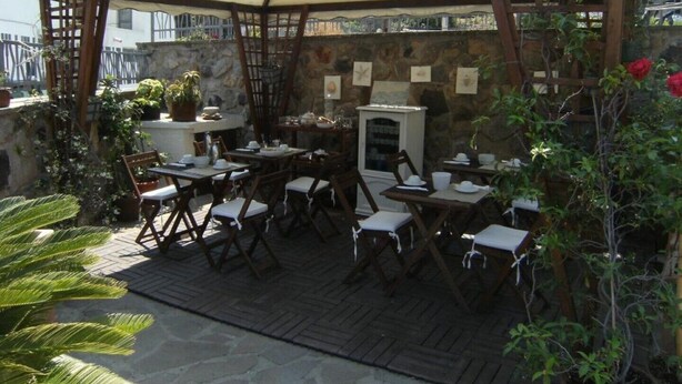 Een terras buiten met houten tafels en stoelen, een witte luifel en een stenen muur.