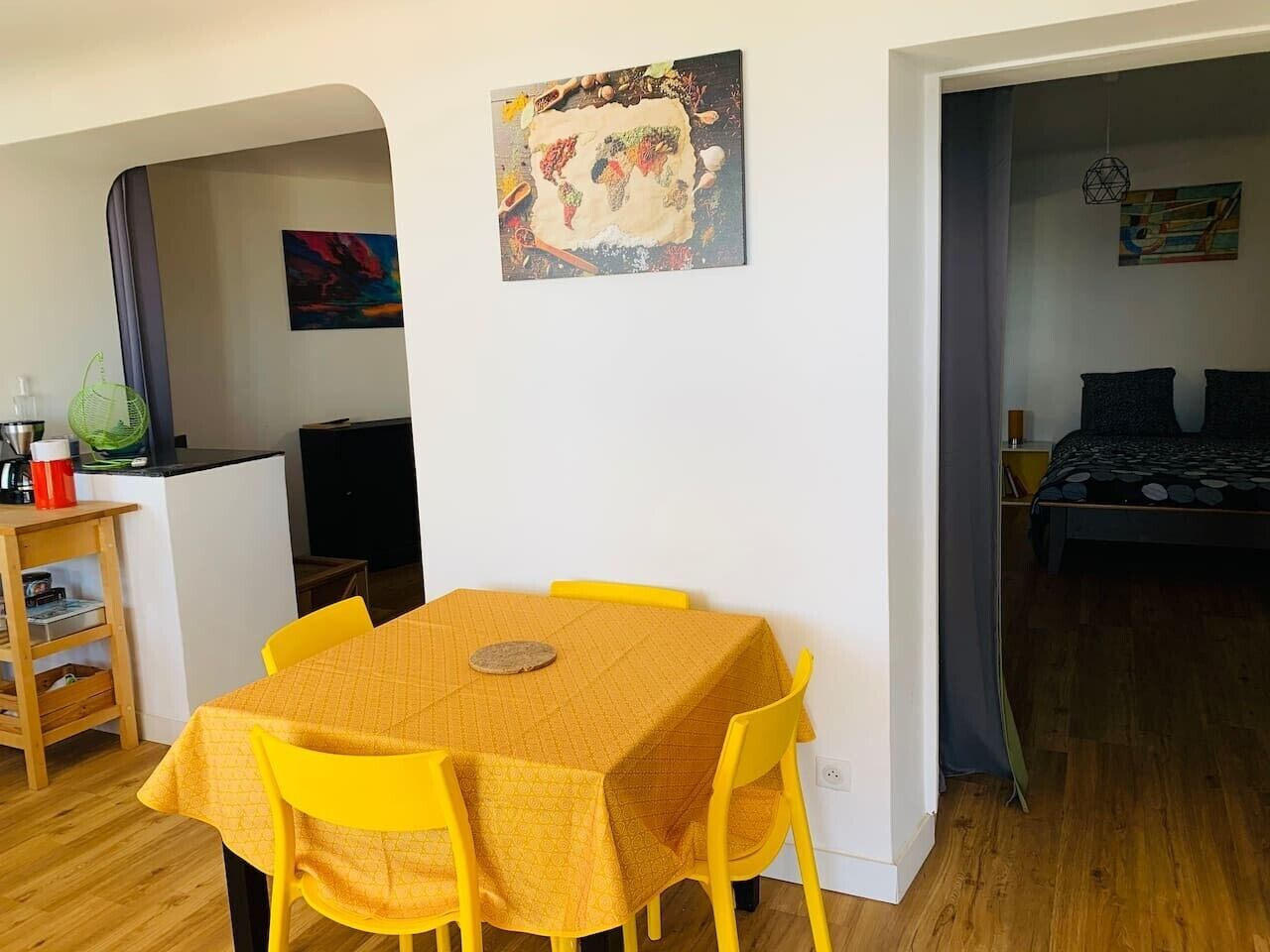 Un coin repas avec une table et des chaises, un coin cuisine avec un évier et des placards, et un lit recouvert d’une couvre-lit de couleur sombre.