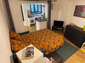Quarto de hotel com cama, uma mesinha, televisão, escrivaninha e cozinha compacta ao fundo.