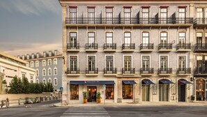 Un edificio de varios pisos con una fila de balcones, un comercio con toldos y un paso de peatones.