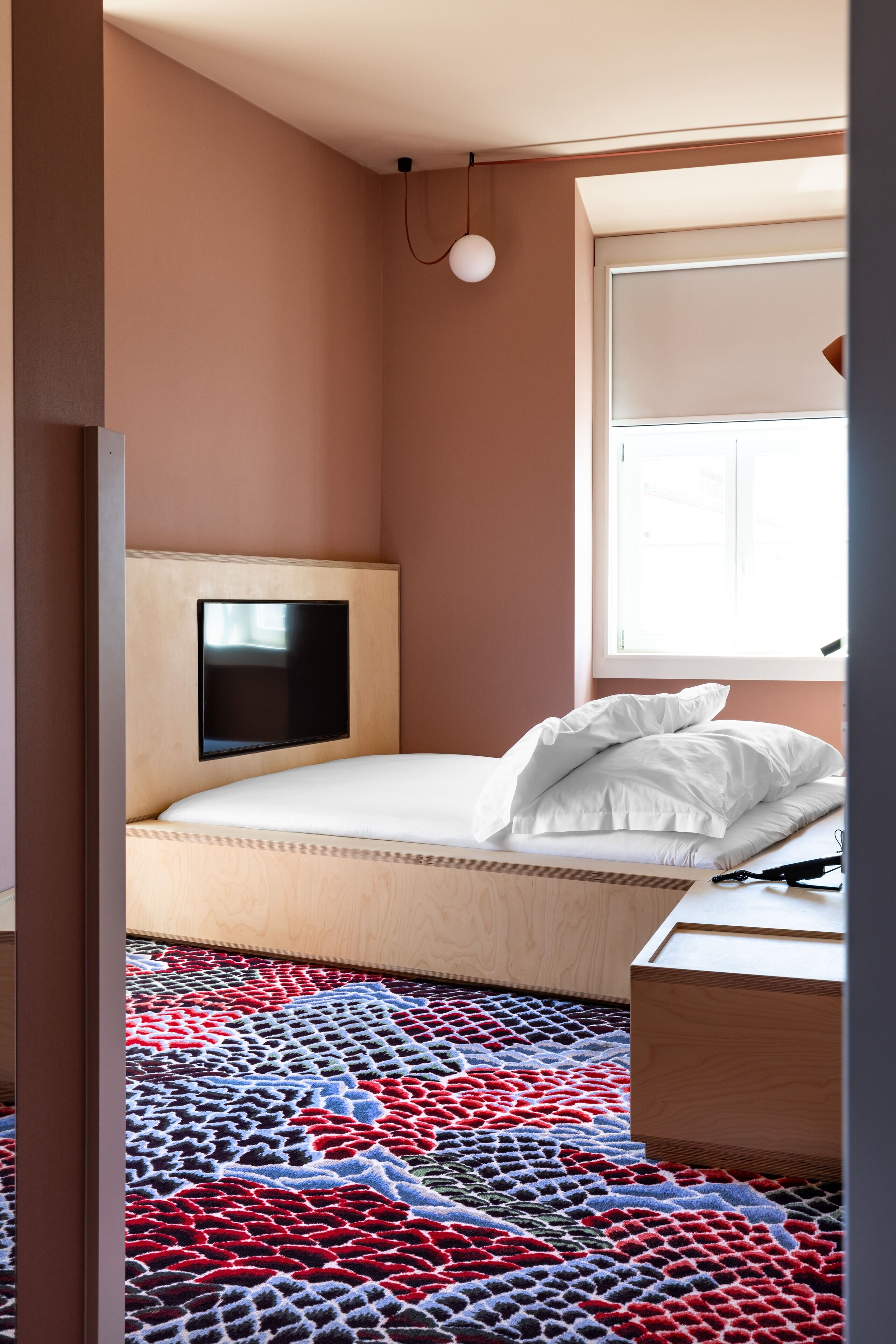 Un dormitorio con cama, televisor montado, ventana con persianas y alfombra estampada.