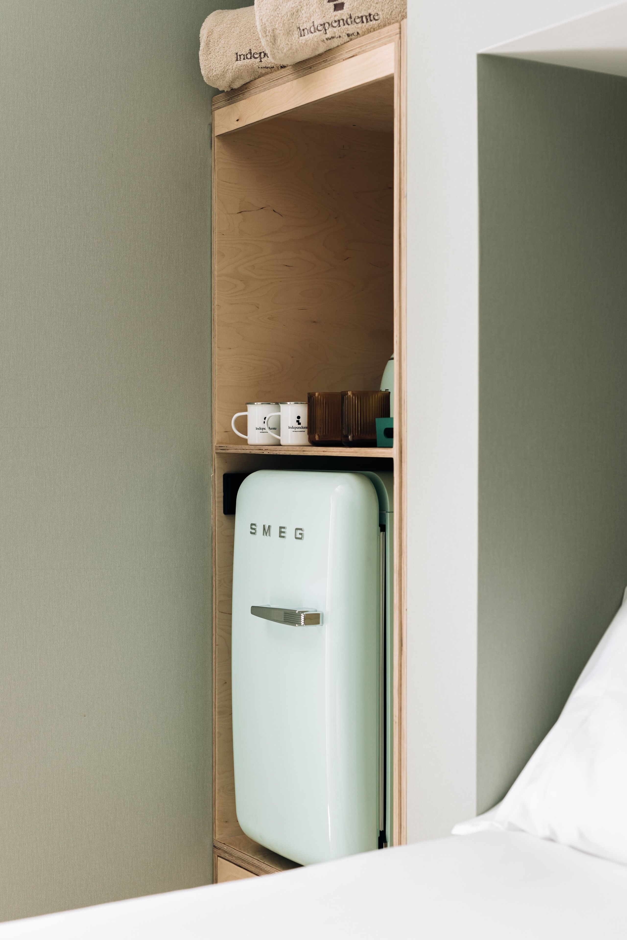 A compact room with a wooden shelf containing a white mug, a small box, and a white pillow. A white Smeg refrigerator is visible in the corner.